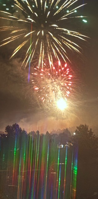 This photo shows three fireworks on top of one another: a silvery firework as it begins to fall, a reddish-white firework in the midst of its burst, and a very bright silvery-white firework just beginning to rise in the night sky.  Below, a diffraction pattern from one of the fireworks almost makes it look like the rainbow effect itself is falling onto the ground.  